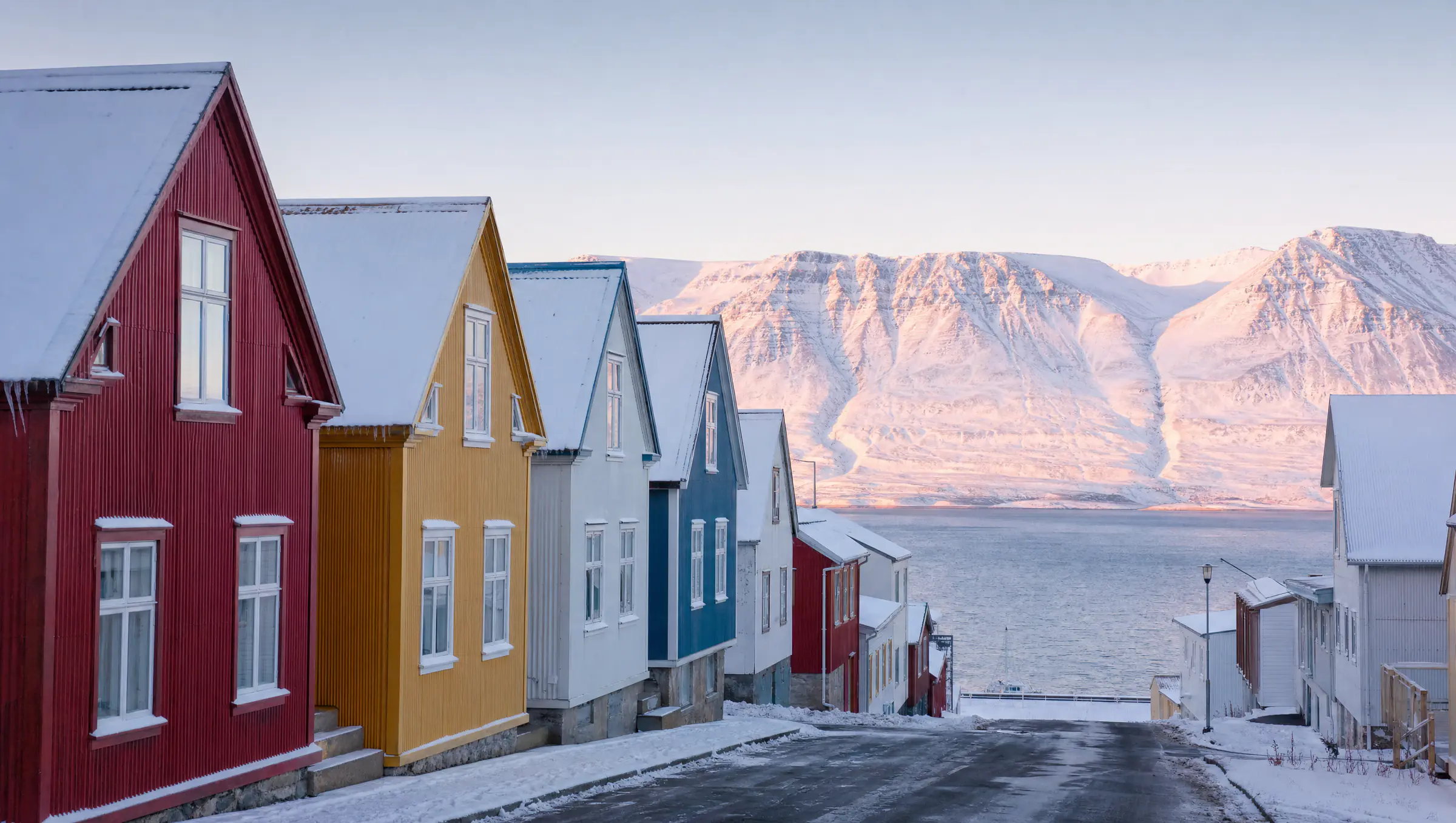 Litrík bárujárnshús í röð við götu sem liggur niður að firði, snævi þakin fjöll handan fjarðarins í vetrarbirtu.